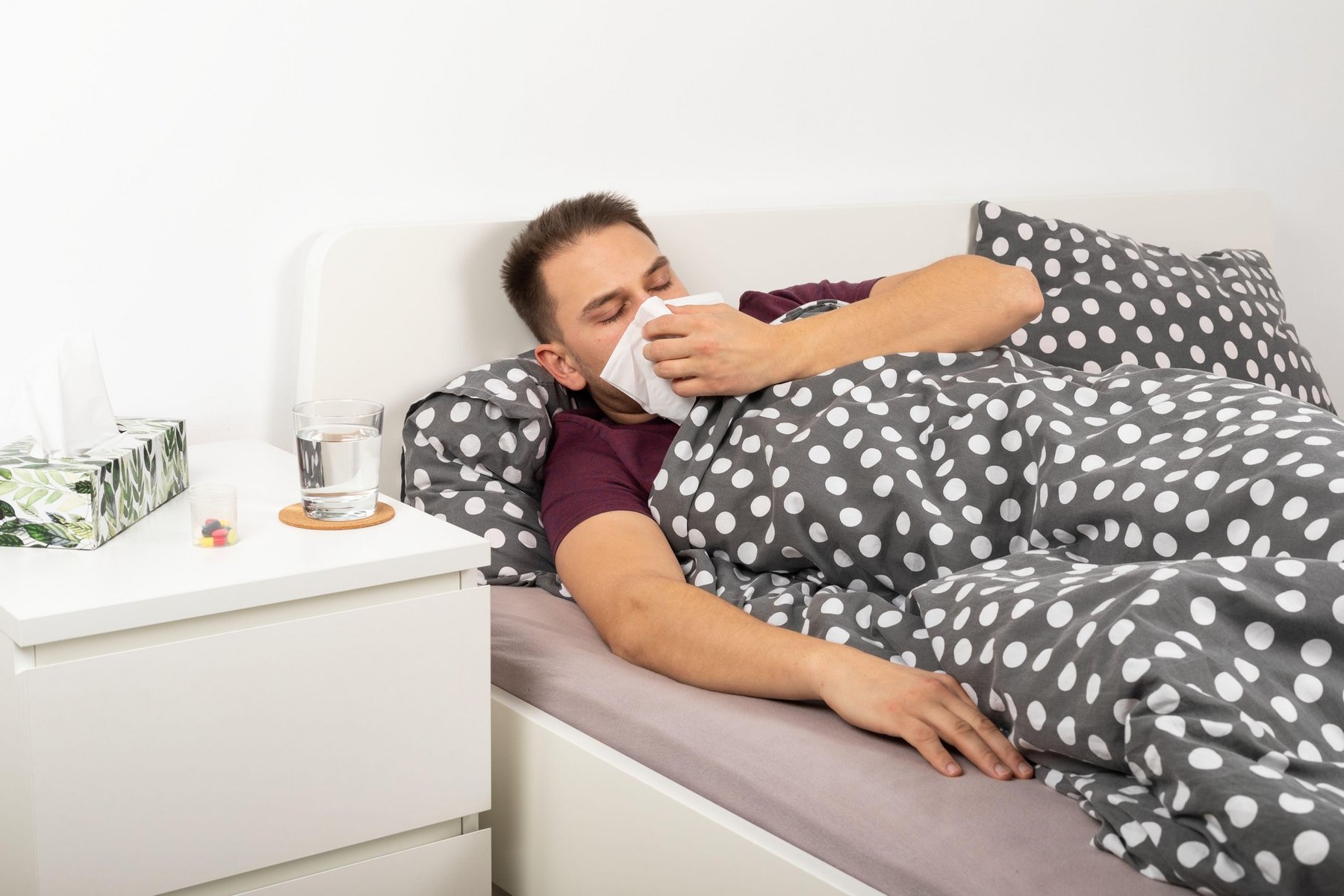 Young man with seasonal influenza is lying in bed at home, blowing his nose in a paper tissue