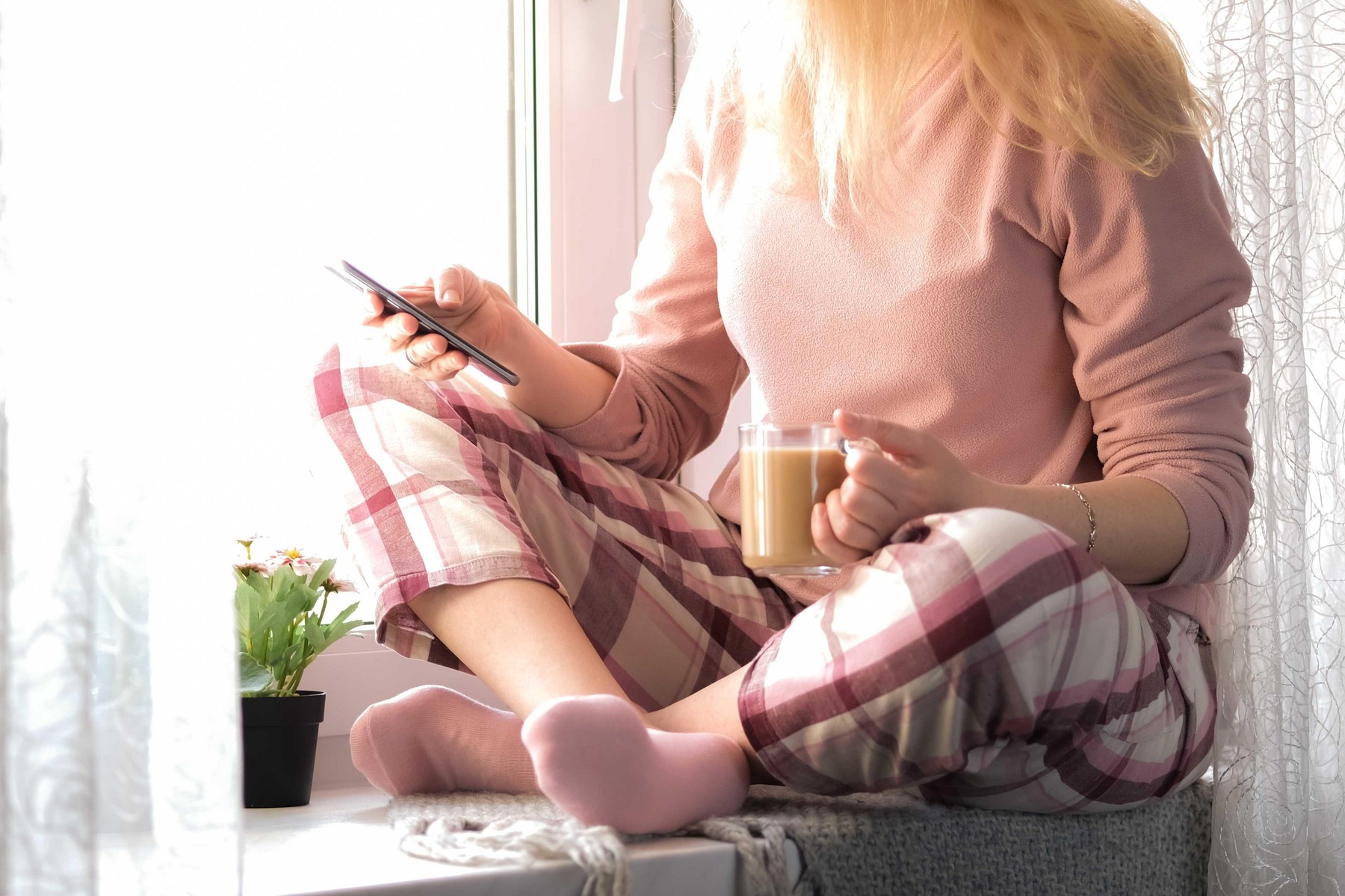 Cropped image of a blonde woman in her pijamas sitting on a window sill with a cup of coffee and her smartphone in hands. Self isolation, stay home or quarantine concept.