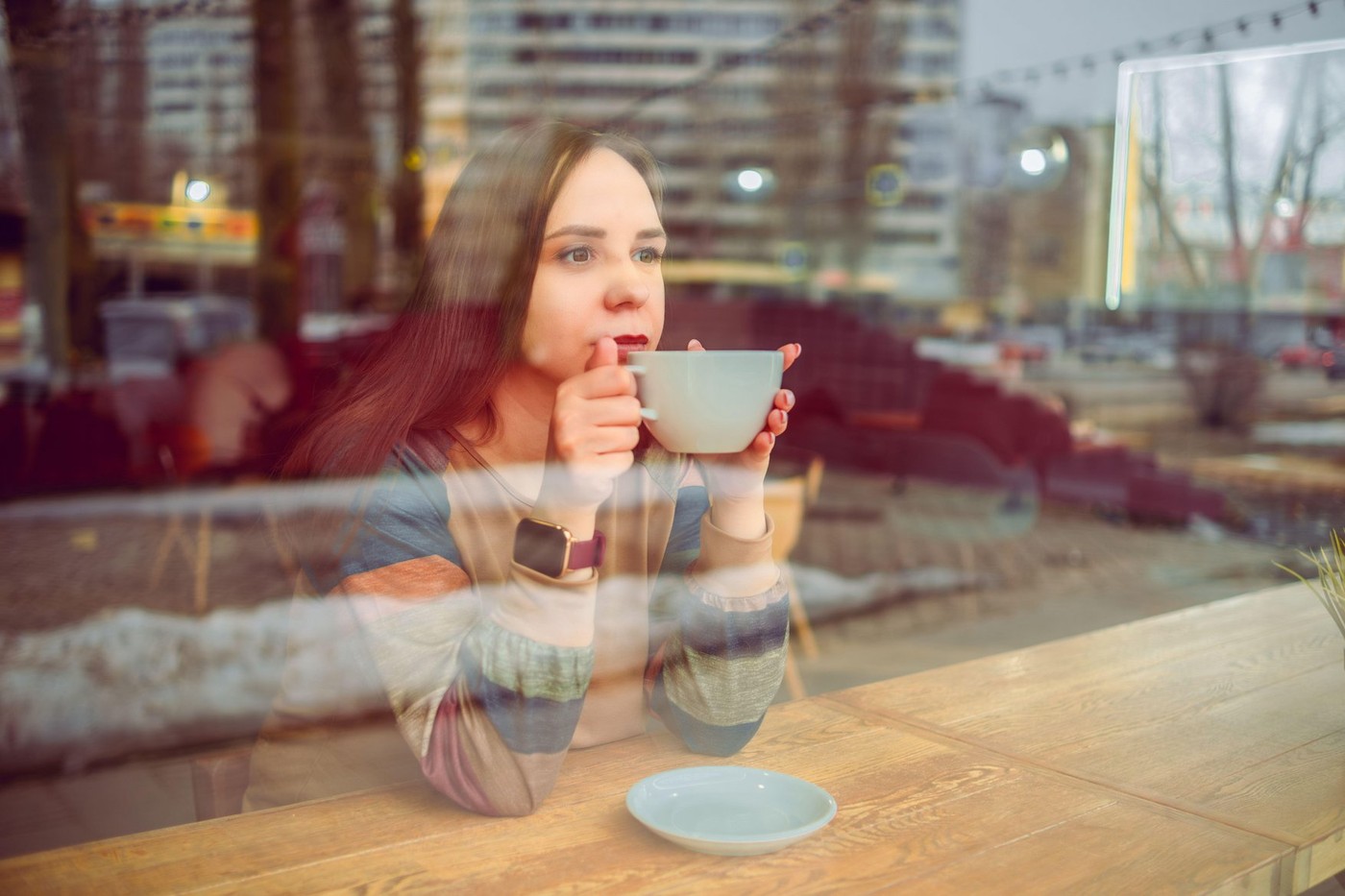 A woman with brunette hair is seated at a cozy cafe table, holding a cup of coffee. She appears relaxed and is enjoying her beverage