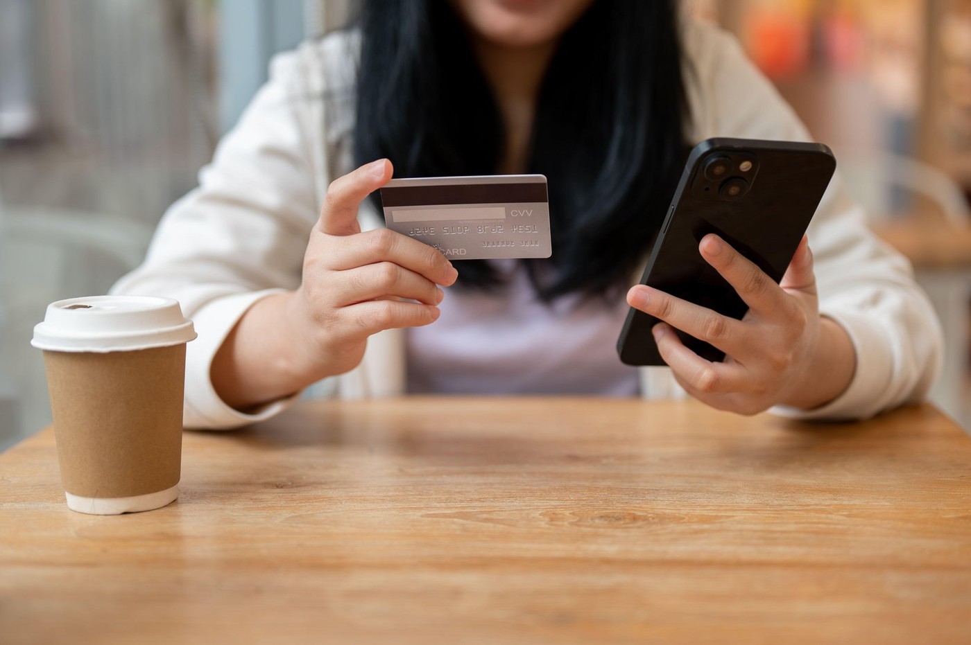 A cropped image of an Asian woman sits at a table indoors using a mobile banking app or shopping online on her smartphone. smartphone and credit card,