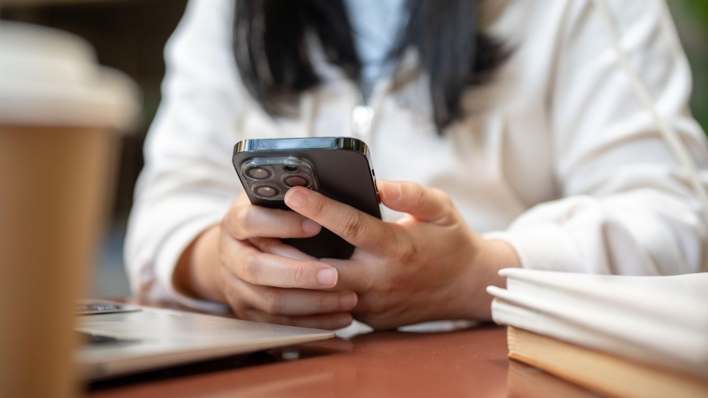 A cropped image of a woman in a white sweater using her smartphone at a table in a cafe, working remotely. scrolling on phone, checking messages, chat