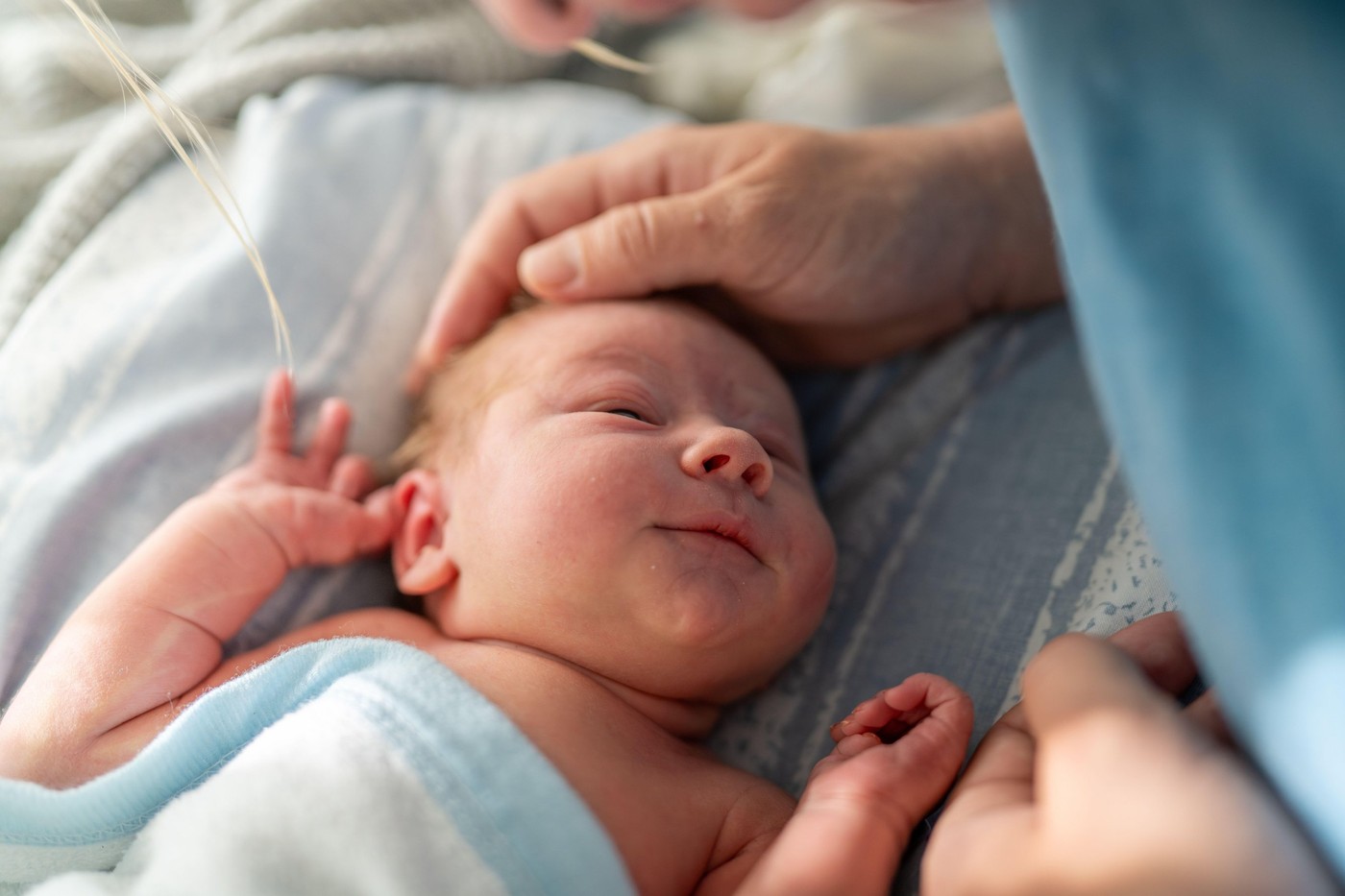 An infant wrapped in a blue blanket, lying down and looking upwards at their mother. The mothers hand gently touches the babys head, capturing a momen