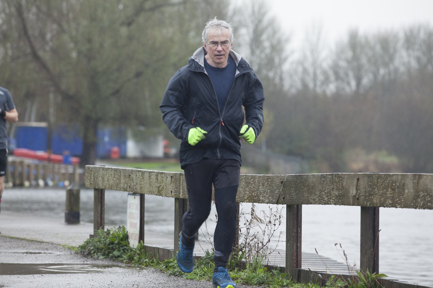 A mature man jogging along a waterside path on an overcast day, wearing a black jacket and bright green gloves.