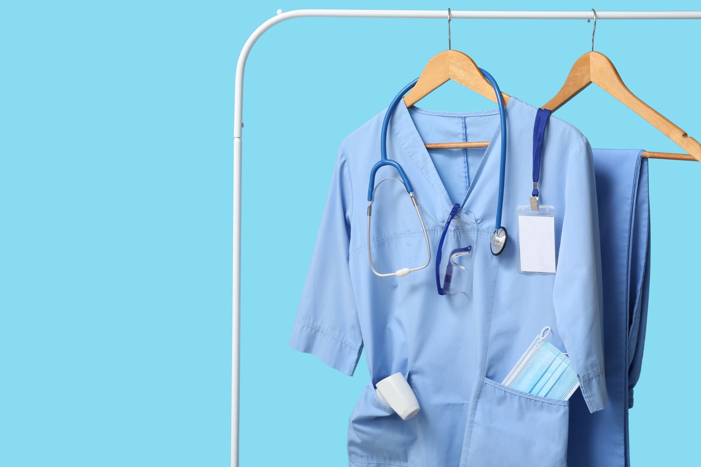 Medical uniform hanging on rack with stethoscope and infrared thermometer on blue background