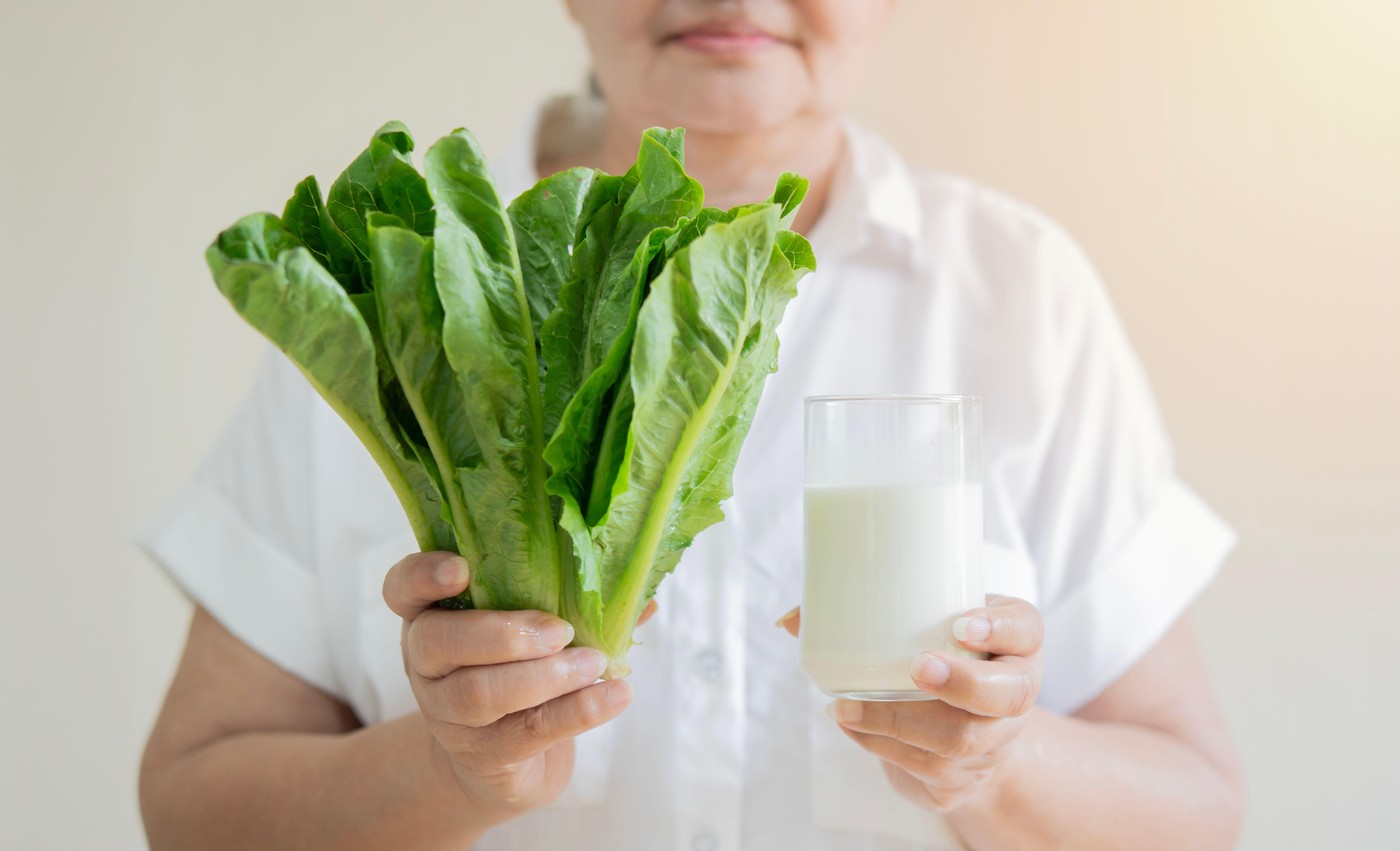 Fresh cos salad and milk glass in hands of smart healthy older woman while standing over light beige background. Selective focus.