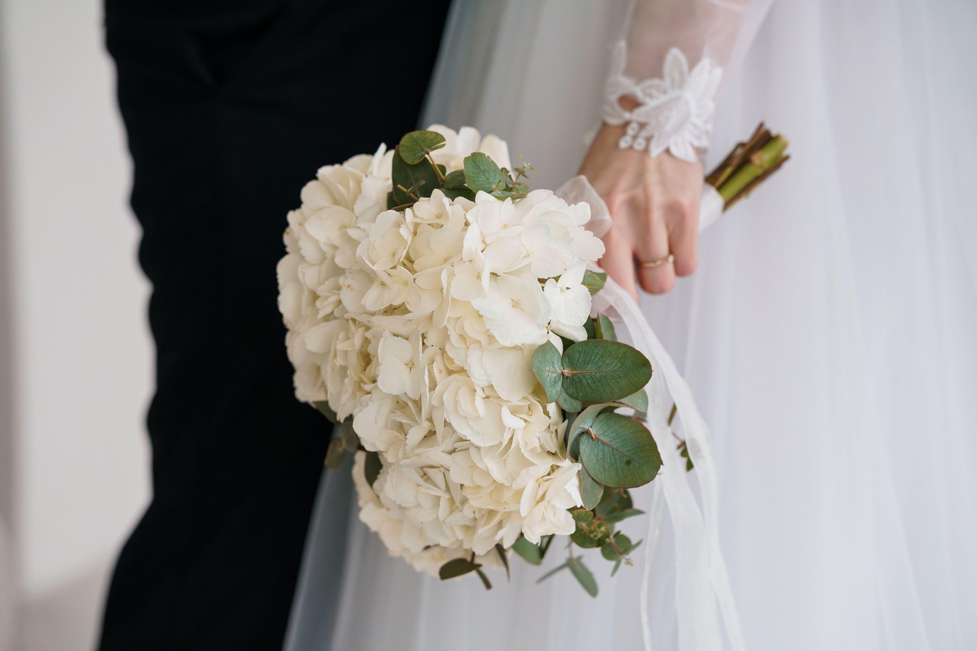 Intimate Bridal Bouquet and Groom's Attire Detail Shot Showcasing White Hydrangeas with Textured Lace Cuff and a Classic Black Suit.