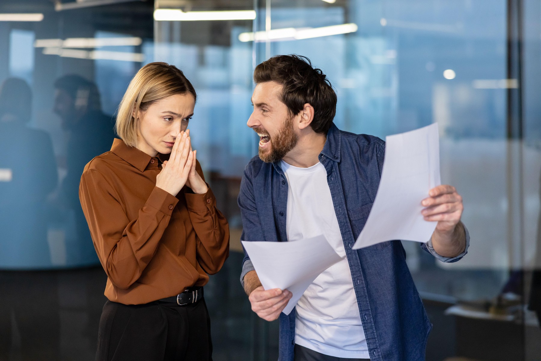 Angry male boss yelling at a stressed female employee holding her hands together.