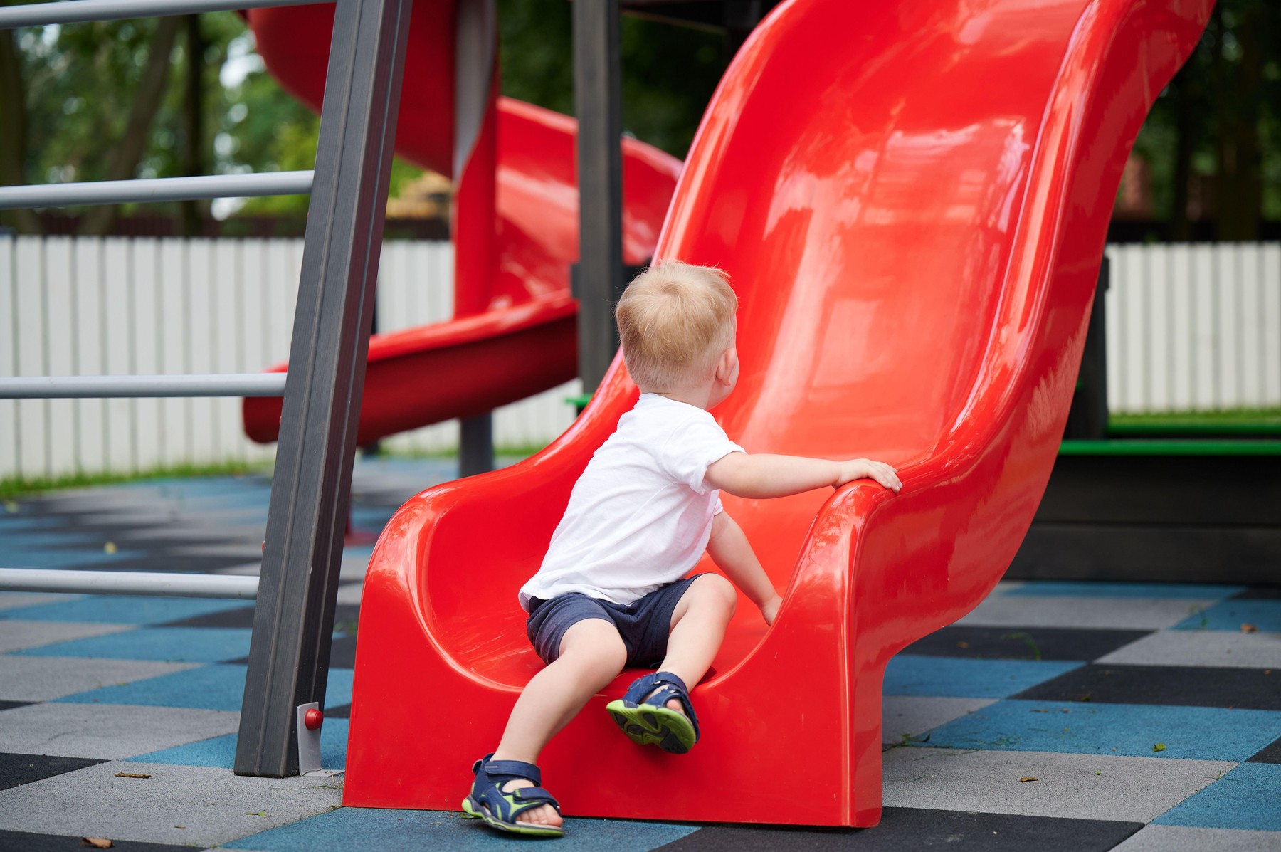 Child sitting on playground slide at bottom, back view, holding side and looking up, summer outfit, bright red slide in park, outdoor activity, active play,Image: 1141656302, License: Royalty-free, Restrictions: , Model Release: yes