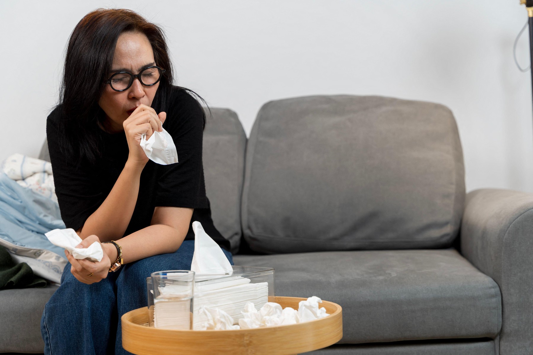 Young woman with cold or flu resting on couch, coughing into tissue and looking tired.
