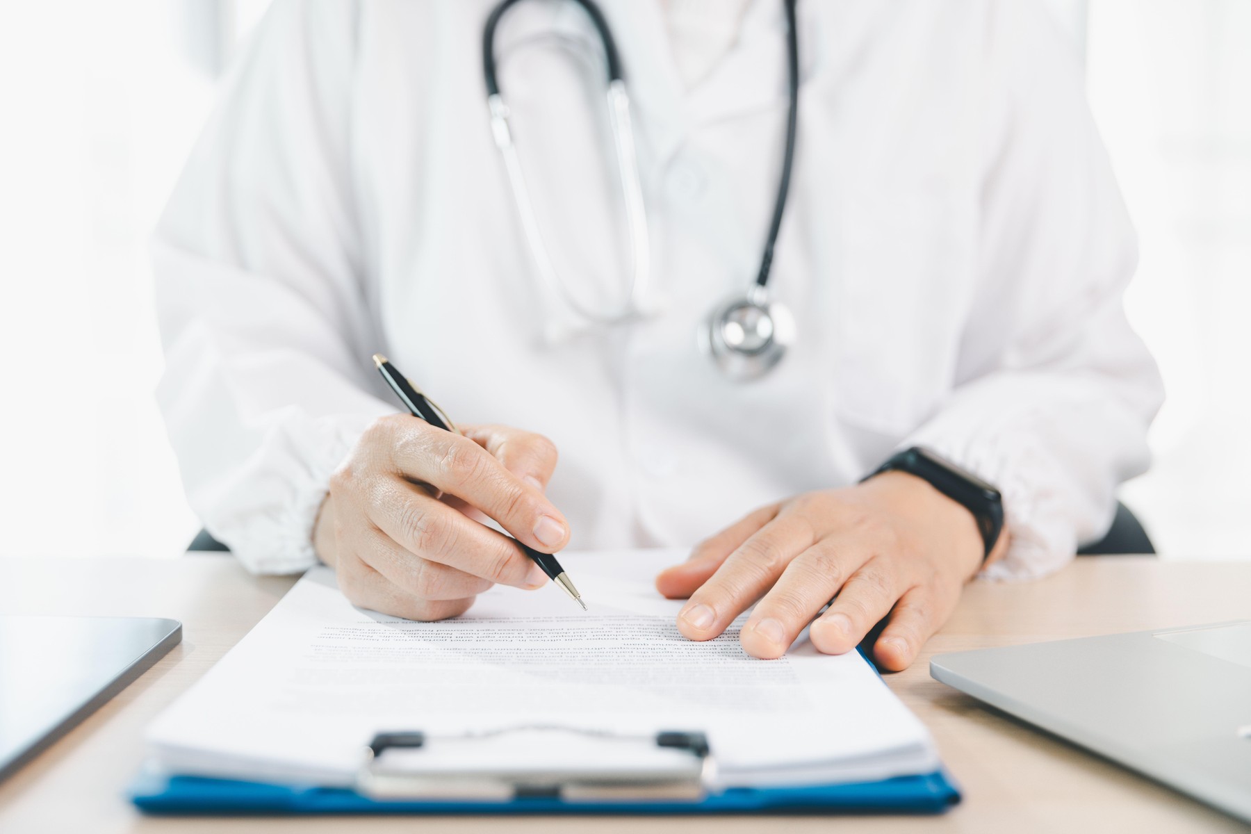 Doctor in white coat signs medical form on clipboard at clinic desk with stethoscope